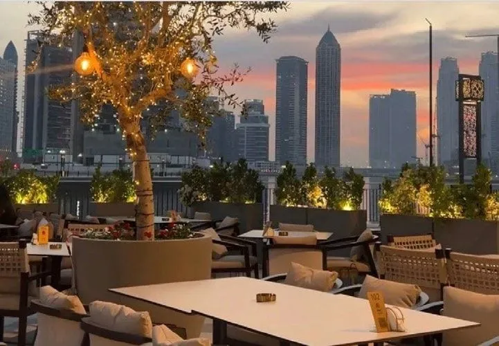 The Dubai terrace at dusk beneath an olive tree hung with lanterns, the towers behind